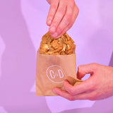 Hand holding a White Chocolate & Pretzel New York Cookie in a Crumbs & Doilies branded paper bag against a purple background